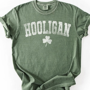 May include: Olive green t-shirt with the word "HOOLIGAN" in distressed white lettering and a white shamrock graphic. The shirt has a crew neck and rolled-up sleeves. The fabric appears soft and slightly faded.
