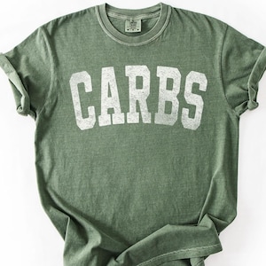 May include: Olive green t-shirt with the word "CARBS" printed in large, distressed white letters. The shirt has a crew neck and rolled-up sleeves. The fabric appears soft and slightly faded, giving it a vintage look.