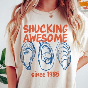 May include: A cream colored t-shirt with the text "Shucking Awesome since 1985" and three blue and white oyster illustrations.