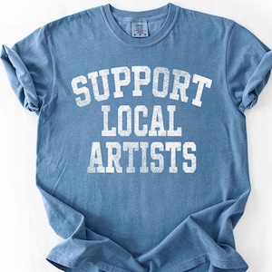 May include: A blue t-shirt with the text "SUPPORT LOCAL ARTISTS" in white block letters. The shirt has a crew neck and short sleeves, and is made of a soft, comfortable material. The shirt is folded on a white surface.