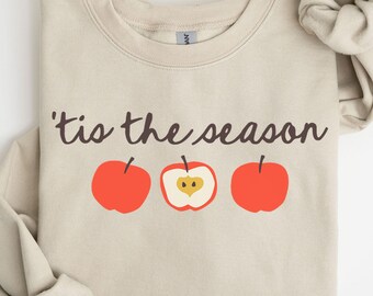 May include: Sand-colored sweatshirt with the phrase "'tis the season" in a cursive font. Below the text are three red apple illustrations, one cut in half to show the seeds. The sleeves are rolled up.