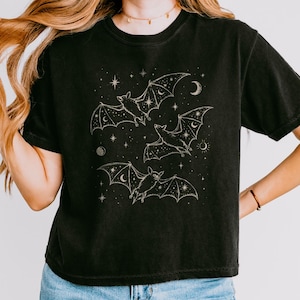 May include: A black short-sleeved t-shirt featuring a graphic print of three flying bats surrounded by celestial elements including stars, crescents, and circular moons. The design is rendered in a light, thin-lined, off-white ink style.
