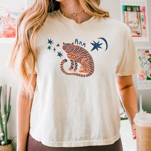 May include: Ivory-colored t-shirt featuring a seated tiger illustration in orange and navy blue, with the word "RAA" above the tiger. The design includes stars and a crescent moon. The shirt is part of a casual wear collection.