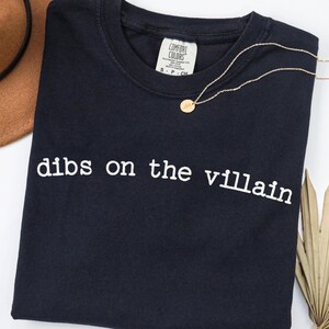 May include: A navy blue t-shirt with the phrase "dibs on the villain" printed in white. A gold necklace with a circular pendant rests on the shirt. A brown hat and dried palm leaf are in the background.