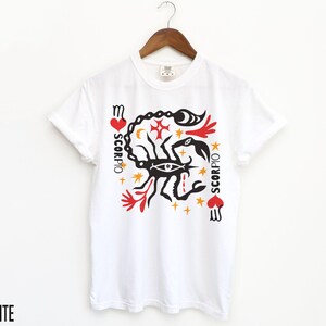 K&ouml;nnte beinhalten: Wei&szlig;es T-Shirt mit einem schwarzen Skorpion-Motiv und dem Wort "SCORPIO" in rot-schwarzer Schrift. Die Grafik enth&auml;lt auch rote Herzen, Sterne und andere Symbole.