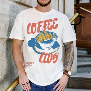 Puede incluir: Camiseta blanca con un gr&aacute;fico de una taza de caf&eacute; con un croissant en la parte superior. El texto "COFFEE CLUB" est&aacute; escrito en una fuente azul y naranja. El texto "EVERYDAY ALL DAY" est&aacute; escrito en una fuente m&aacute;s peque&ntilde;a debajo del gr&aacute;fico.