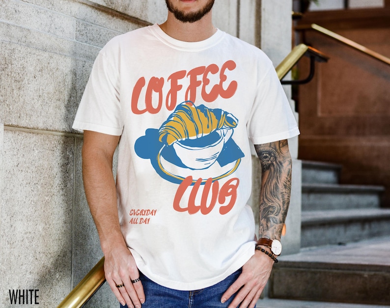 Puede incluir: Camiseta blanca con un gr&aacute;fico de una taza de caf&eacute; con un croissant en la parte superior. El texto "COFFEE CLUB" est&aacute; escrito en una fuente azul y naranja. El texto "EVERYDAY ALL DAY" est&aacute; escrito en una fuente m&aacute;s peque&ntilde;a debajo del gr&aacute;fico.