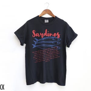 May include: Black t-shirt with a red and blue graphic of sardines and a text description about the fish. The text reads "Sardines" in red and the rest of the text is in blue.