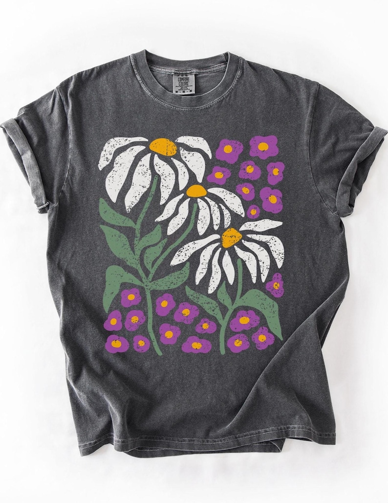 May include: Dark gray t-shirt featuring a floral design. The graphic includes large white daisies with yellow centers, green stems and leaves, and small purple flowers. The shirt has a crew neck and rolled-up sleeves.