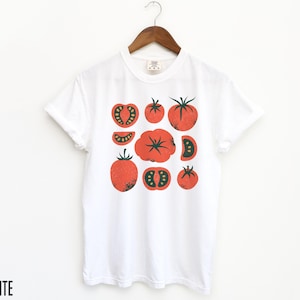 May include: White t-shirt featuring a graphic print of red tomatoes in various shapes and sizes. The tomatoes have green stems and leaves.