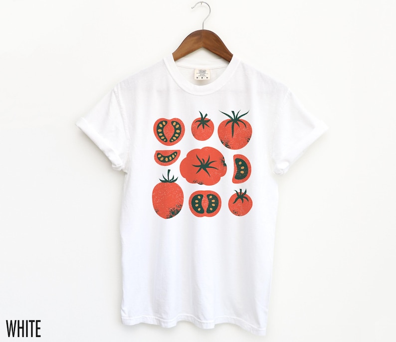 May include: White t-shirt featuring a graphic print of red tomatoes in various shapes and sizes. The tomatoes have green stems and leaves.