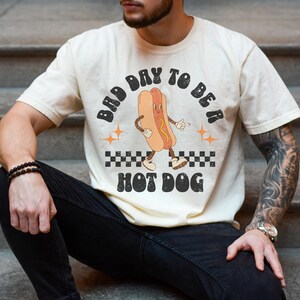 May include: A person wearing a cream colored t-shirt with a retro graphic of a hot dog with the text "Bad Day To Be A Hot Dog".