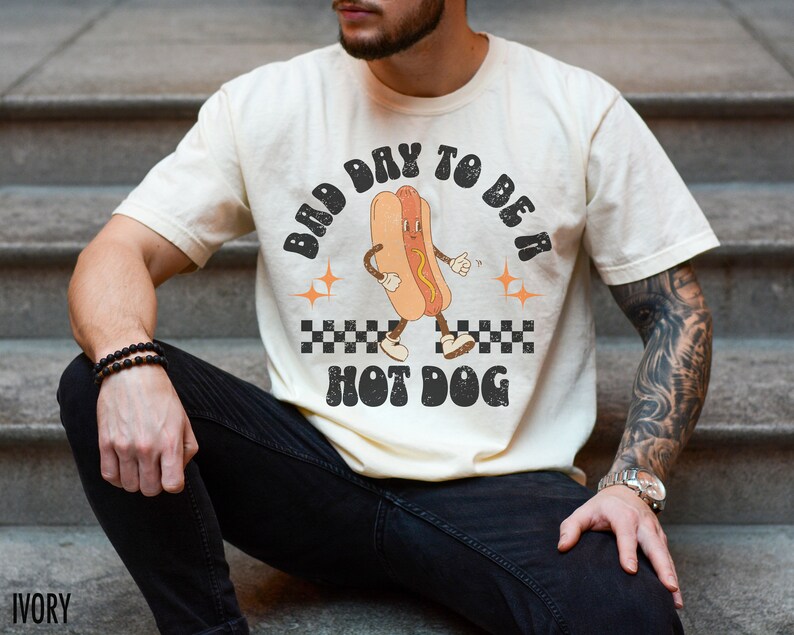 May include: A person wearing a cream colored t-shirt with a retro graphic of a hot dog with the text "Bad Day To Be A Hot Dog".
