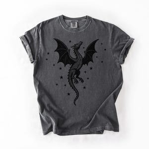 Op de afbeelding: Donkergrijze t-shirt met een zwart drakenontwerp met uitgespreide vleugels en sterren. De draak is gedetailleerd. Het shirt heeft een ronde hals en korte mouwen.