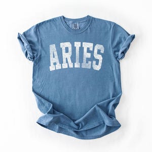 May include: A blue t-shirt with the word "ARIES" printed in white, featuring a vintage, distressed design. The shirt is made of soft material and has rolled-up sleeves.