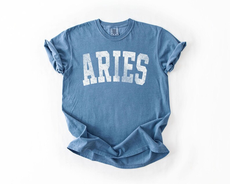 May include: A blue t-shirt with the word "ARIES" printed in white, featuring a vintage, distressed design. The shirt is made of soft material and has rolled-up sleeves.