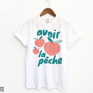 May include: White t-shirt with a graphic of peaches and the text "avoir la p&ecirc;che" in teal.
