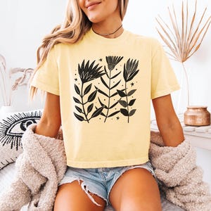 May include: A pale yellow t-shirt with a black floral print. The design features stylized flowers, leaves, and stars. The short-sleeved shirt is made of a soft material.