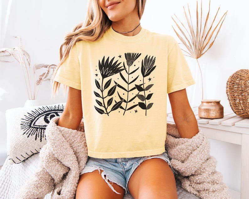 May include: A pale yellow t-shirt with a black floral print. The design features stylized flowers, leaves, and stars. The short-sleeved shirt is made of a soft material.