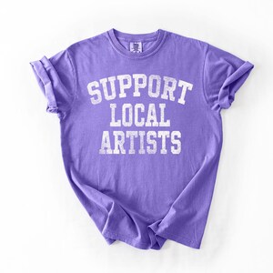 May include: A lavender t-shirt with rolled-up sleeves. The shirt features the text "SUPPORT LOCAL ARTISTS" in a distressed, white, collegiate-style font. The shirt is made of a soft material.