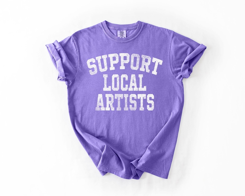 May include: A lavender t-shirt with rolled-up sleeves. The shirt features the text "SUPPORT LOCAL ARTISTS" in a distressed, white, collegiate-style font. The shirt is made of a soft material.