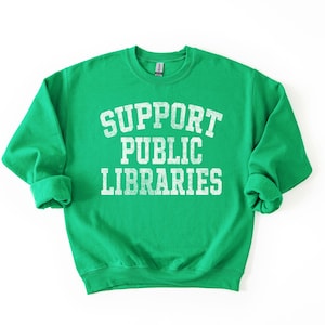 May include: A bright green sweatshirt with the text "SUPPORT PUBLIC LIBRARIES" in distressed white lettering. The crew neck sweatshirt has long sleeves with rolled cuffs. The design is centered on the front of the garment.