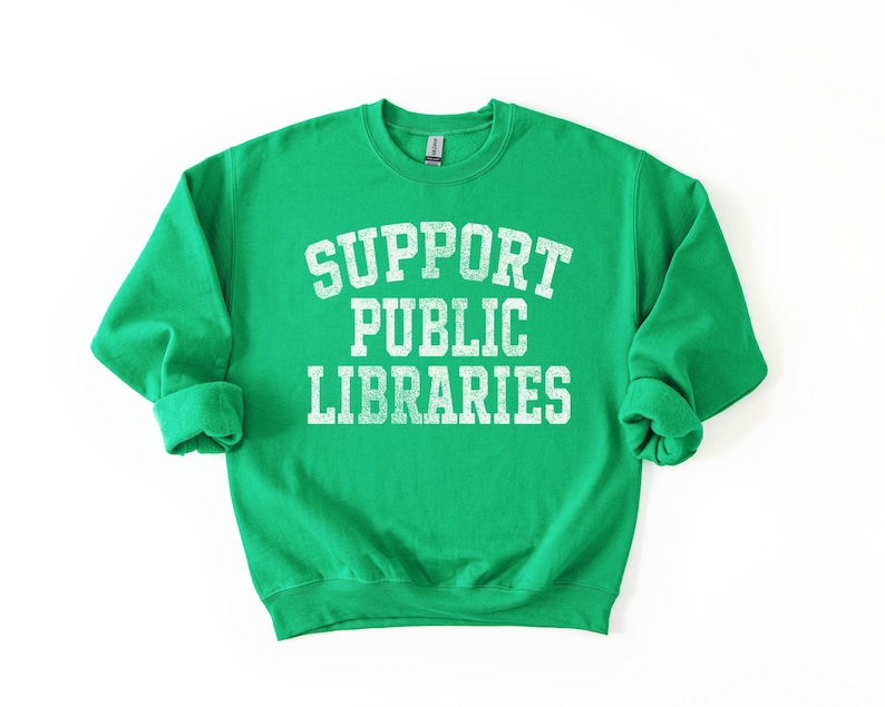 May include: A bright green sweatshirt with the text "SUPPORT PUBLIC LIBRARIES" in distressed white lettering. The crew neck sweatshirt has long sleeves with rolled cuffs. The design is centered on the front of the garment.