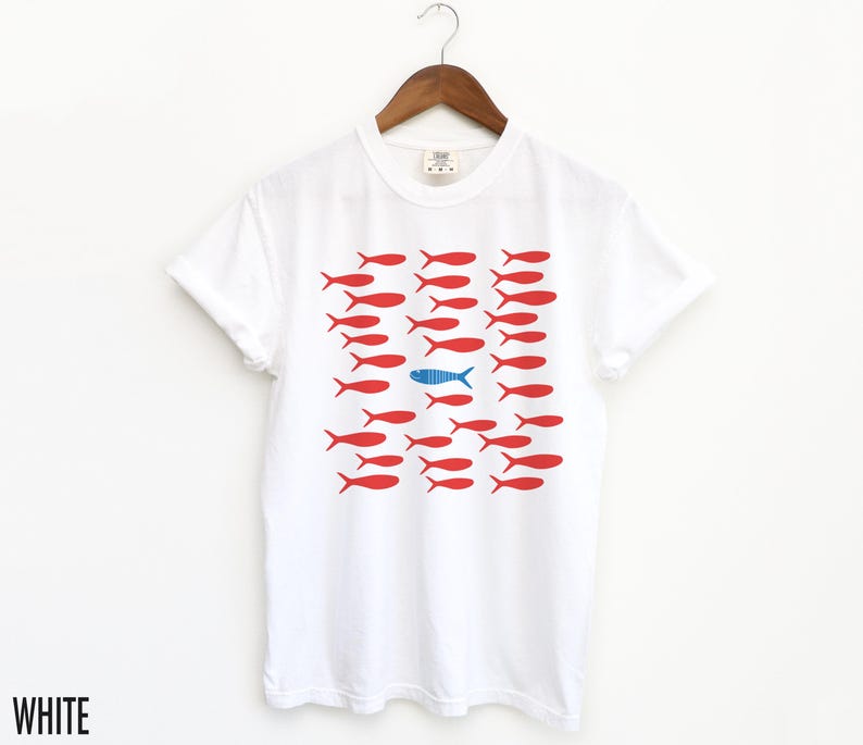 May include: White t-shirt with a graphic of red fish swimming in a row, with one blue fish swimming in the opposite direction. The text "WHITE" is visible at the bottom of the image.