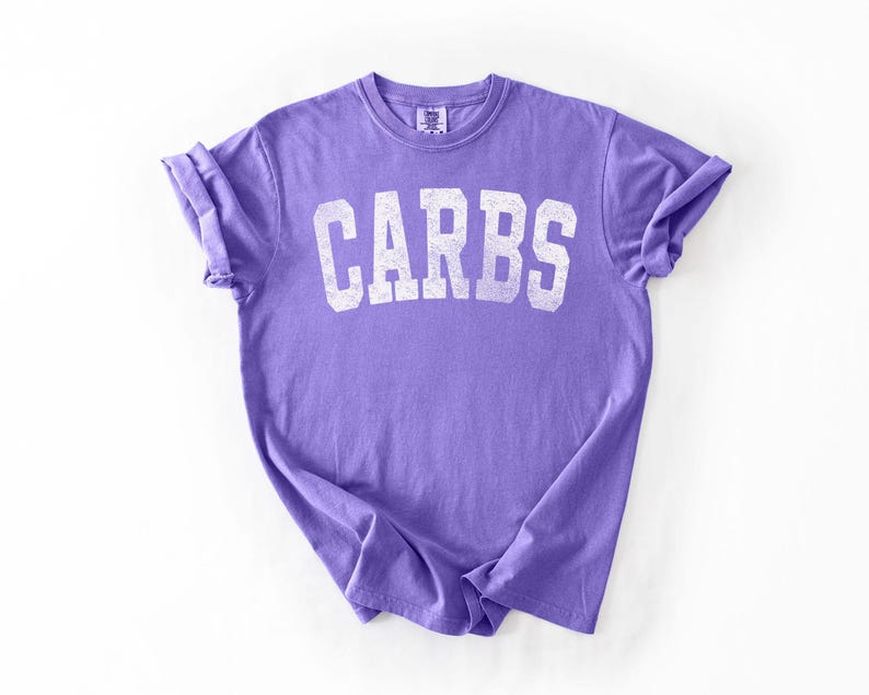 May include: A purple t-shirt with the word "CARBS" printed in white, arched lettering. The shirt has rolled-up sleeves and a crew neck. The fabric appears soft and slightly faded.