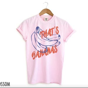 Puede incluir: Una camiseta rosa claro con un gr&aacute;fico de pl&aacute;tanos y el texto "That's Bananas" en naranja y azul. La camiseta est&aacute; colgada en una percha de madera.
