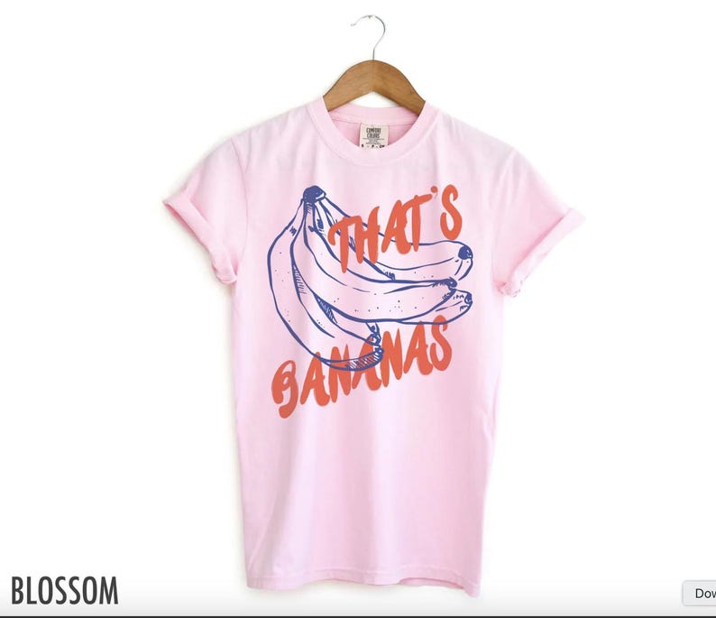 Puede incluir: Una camiseta rosa claro con un gr&aacute;fico de pl&aacute;tanos y el texto "That's Bananas" en naranja y azul. La camiseta est&aacute; colgada en una percha de madera.
