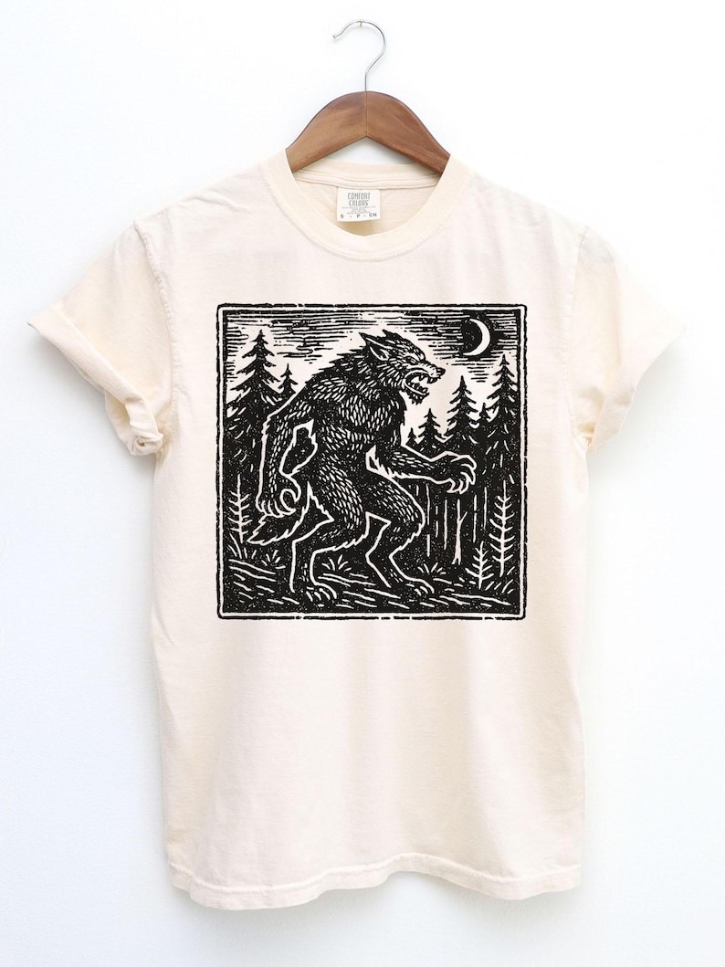 Puede incluir: Camiseta color crema con una ilustraci&oacute;n en blanco y negro de un hombre lobo en un bosque bajo una luna creciente. El dise&ntilde;o est&aacute; en un marco cuadrado, con un estilo de grabado en madera vintage. La camiseta est&aacute; colgada en una percha de madera.