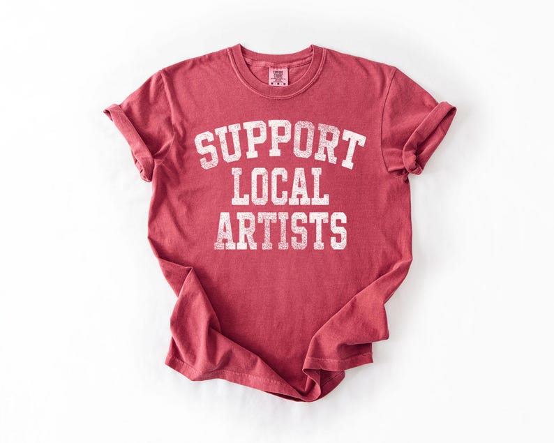 May include: A red t-shirt with rolled-up sleeves. The shirt features the text "SUPPORT LOCAL ARTISTS" in a distressed, white, collegiate-style font. The shirt is made of a soft material.