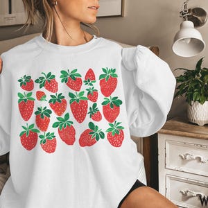 K&ouml;nnte beinhalten: Wei&szlig;er Pullover mit einem grafischen Druck von roten Erdbeeren auf wei&szlig;em Hintergrund. Der Pullover hat einen Rundhalsausschnitt und lange &Auml;rmel.