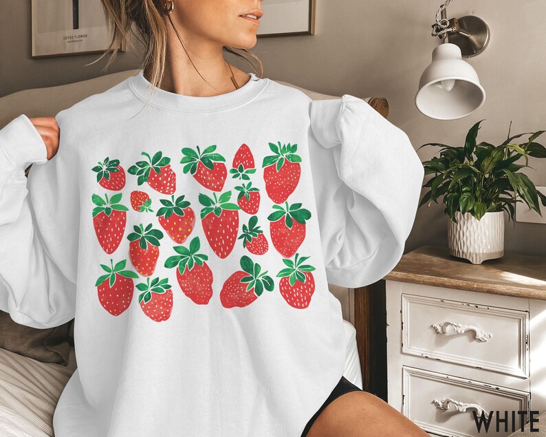K&ouml;nnte beinhalten: Wei&szlig;er Pullover mit einem grafischen Druck von roten Erdbeeren auf wei&szlig;em Hintergrund. Der Pullover hat einen Rundhalsausschnitt und lange &Auml;rmel.