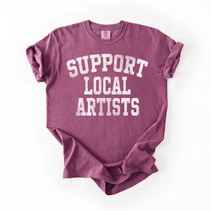 May include: A maroon t-shirt with the text "SUPPORT LOCAL ARTISTS" in distressed white lettering. The shirt has short sleeves and a crew neck. The fabric appears soft and comfortable.