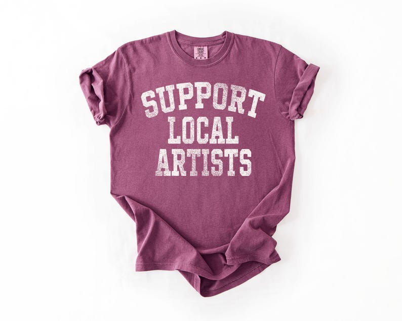 May include: A maroon t-shirt with the text "SUPPORT LOCAL ARTISTS" in distressed white lettering. The shirt has short sleeves and a crew neck. The fabric appears soft and comfortable.