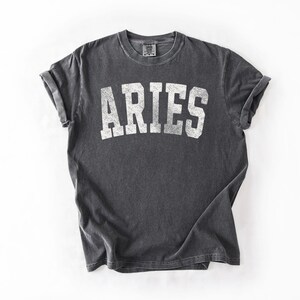 May include: A charcoal gray t-shirt with the word "ARIES" printed in white, block letters. The shirt has rolled-up sleeves and a vintage, worn look. The t-shirt is laid flat on a white surface.