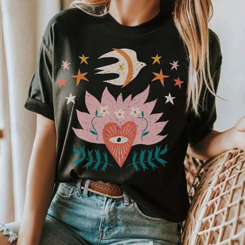May include: Black t-shirt with a distressed graphic. The design features a white bird, pink lotus flower, a heart with an eye, and stars in orange, pink, and yellow. The shirt is made of a soft material.