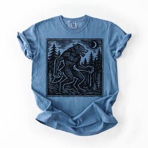 Puede incluir: Camiseta azul con una ilustraci&oacute;n en blanco y negro de un hombre lobo en un bosque bajo una luna creciente. El gr&aacute;fico est&aacute; en un marco rectangular. La camiseta tiene las mangas enrolladas.