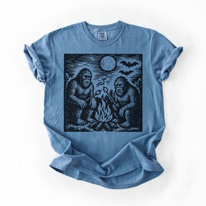May include: A blue t-shirt featuring a black and white graphic of two Bigfoot creatures roasting marshmallows over a campfire under a full moon. Bats are flying in the sky above the moon. The shirt is folded.