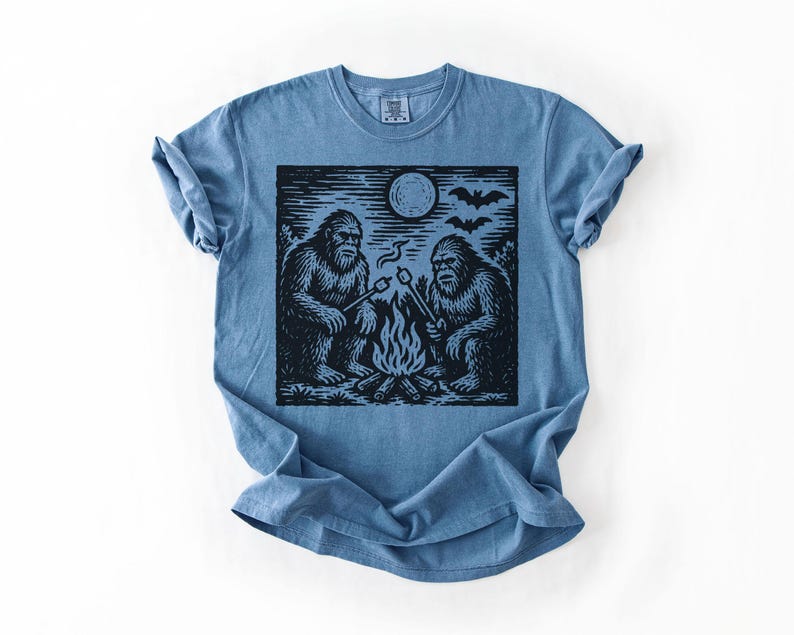 May include: A blue t-shirt featuring a black and white graphic of two Bigfoot creatures roasting marshmallows over a campfire under a full moon. Bats are flying in the sky above the moon. The shirt is folded.