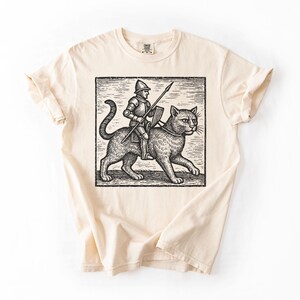 Puede incluir: Camiseta color crema con una ilustraci&oacute;n en blanco y negro de un caballero montando un gato grande. El caballero sostiene una lanza. El gr&aacute;fico est&aacute; en un marco rectangular.