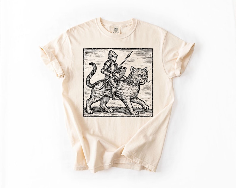 Puede incluir: Camiseta color crema con una ilustraci&oacute;n en blanco y negro de un caballero montando un gato grande. El caballero sostiene una lanza. El gr&aacute;fico est&aacute; en un marco rectangular.