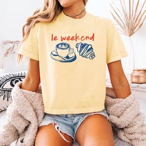 May include: A light yellow t-shirt with the words "le weekend" in red, along with a blue and white coffee cup and croissant graphic. The shirt is a casual style.