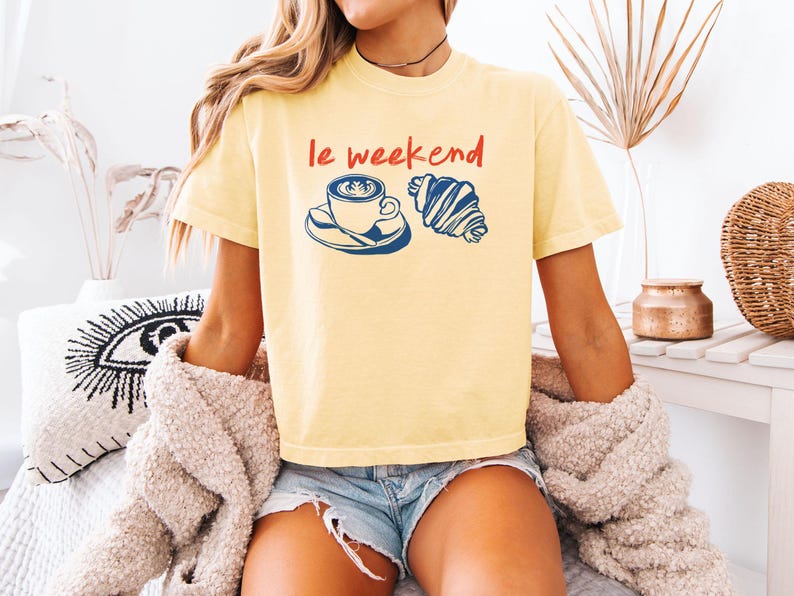 May include: A light yellow t-shirt with the words "le weekend" in red, along with a blue and white coffee cup and croissant graphic. The shirt is a casual style.