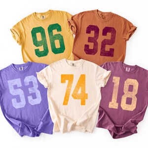 May include: Five short-sleeved t-shirts in various colors: yellow, brown, purple, cream, and burgundy. Each shirt features a large number in a contrasting color, including 96, 32, 53, 74, and 18. The shirts have a vintage, distressed look.