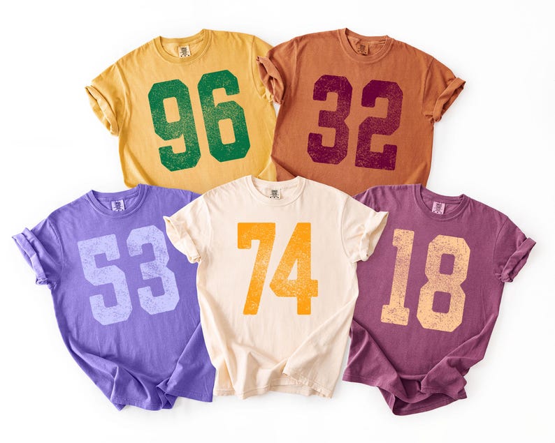 May include: Five short-sleeved t-shirts in various colors: yellow, brown, purple, cream, and burgundy. Each shirt features a large number in a contrasting color, including 96, 32, 53, 74, and 18. The shirts have a vintage, distressed look.