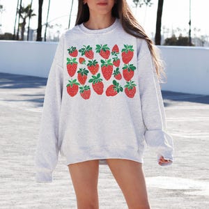K&ouml;nnte beinhalten: Ein grauer Pullover mit einem Grafikdruck von roten Erdbeeren mit gr&uuml;nen Bl&auml;ttern. Der Pullover hat lange &Auml;rmel und einen Rundhalsausschnitt.
