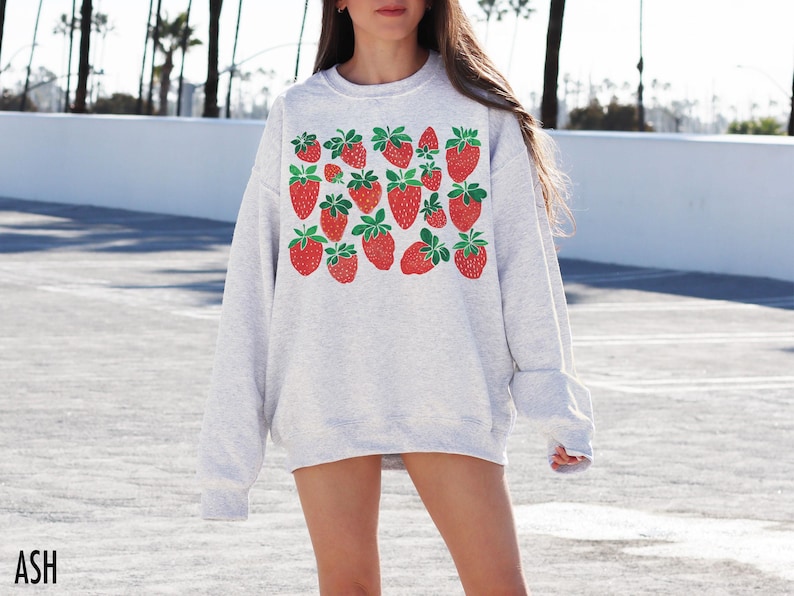 K&ouml;nnte beinhalten: Ein grauer Pullover mit einem Grafikdruck von roten Erdbeeren mit gr&uuml;nen Bl&auml;ttern. Der Pullover hat lange &Auml;rmel und einen Rundhalsausschnitt.